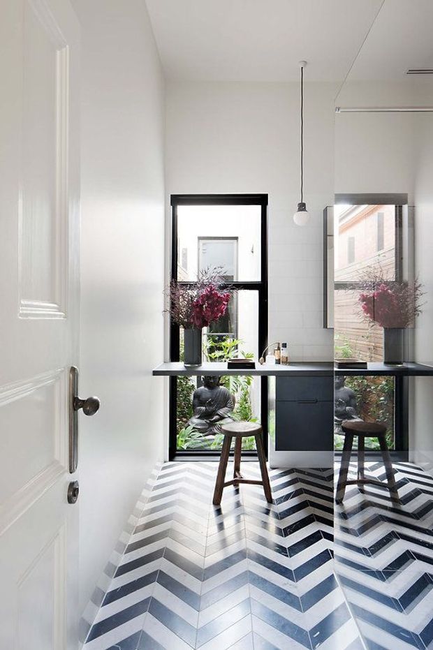 A bright, narrow bathroom viewed from the doorway, featuring a bold black-and-white chevron tile floor, white wall panelling, a floating dark vanity with a white benchtop, black-framed windows, a large wall mirror, a pendant light, and a stool. Potted flowers and greenery are visible by the windows, with a garden statue outside.