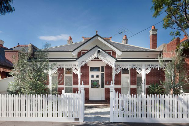 Front exterior view of a single-storey Victorian-era brick cottage with an ornate gabled porch, decorative white timber fretwork, multiple chimneys, tiled pitched roof, and a white picket fence. Mature planting frames the symmetrical facade.