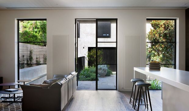 A bright contemporary open-plan living area with a large white kitchen island and bar seating on the right, a grey sectional sofa and low coffee tables on the left, and full-height black-framed glazing and a glass door opening to a planted courtyard. The room has pale timber flooring, light neutral walls, a white paneled ceiling, and slim vertical radiators beside the windows.