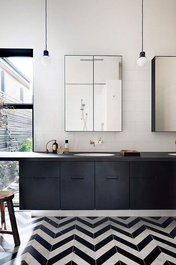 A compact, contemporary bathroom with a long dark floating vanity, integrated basins, frameless mirrored medicine cabinets, white rectangular wall tiles, and exposed pendant lights. A bold black-and-white chevron floor creates a graphic feature, while a large window brings in natural light and views of greenery and neighbouring architecture.