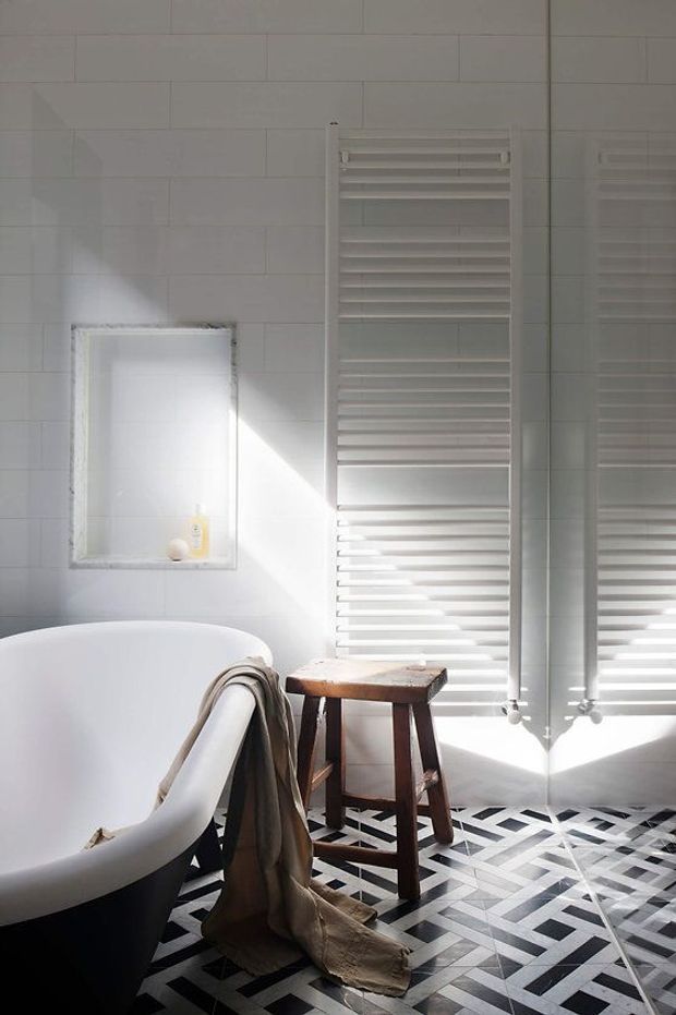 A bright, refined bathroom featuring a freestanding white bathtub with a dark exterior, white rectangular wall tiles, a recessed wall niche, and a tall white heated towel rail. A timber stool and draped towel sit beside the bath. The floor has a bold geometric black-and-white patterned tile design, with strong sunlight entering through a large reflective glazed opening on the right.