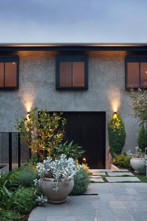 A dusk view of a contemporary residential facade with a textured grey rendered wall, dark-framed upper windows, a black garage or entry door, and warm wall-mounted uplighting. A landscaped approach features large grey paving slabs, planted garden beds, clipped shrubs, ornamental trees, and large ceramic planters.