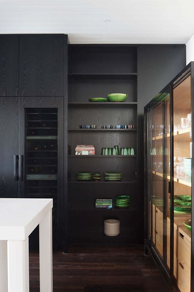 A refined kitchen storage vignette featuring tall dark timber cabinetry, an integrated wine refrigerator, open display shelving with green glassware and ceramics, and a glazed display cabinet with warm timber interiors. A white island edge is visible in the foreground, with dark timber flooring and a white paneled ceiling completing the space.