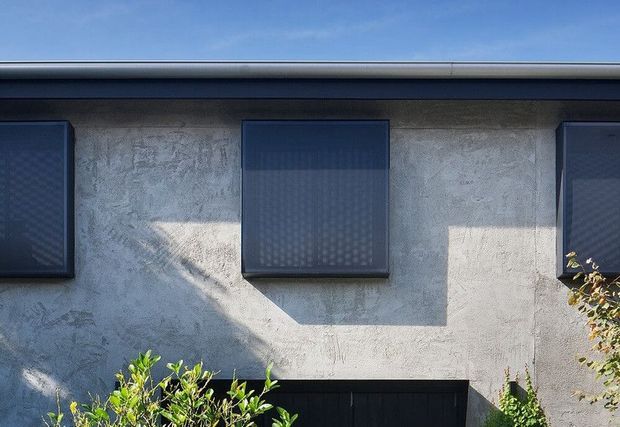 Close-up exterior view of a contemporary single-storey facade with pale textured rendered walls, dark projecting rectangular window surrounds or shutters, and a dark garage or entry opening at the base. Strong sunlight creates angular shadows across the facade, with sparse planting at the edges and clear blue sky above.