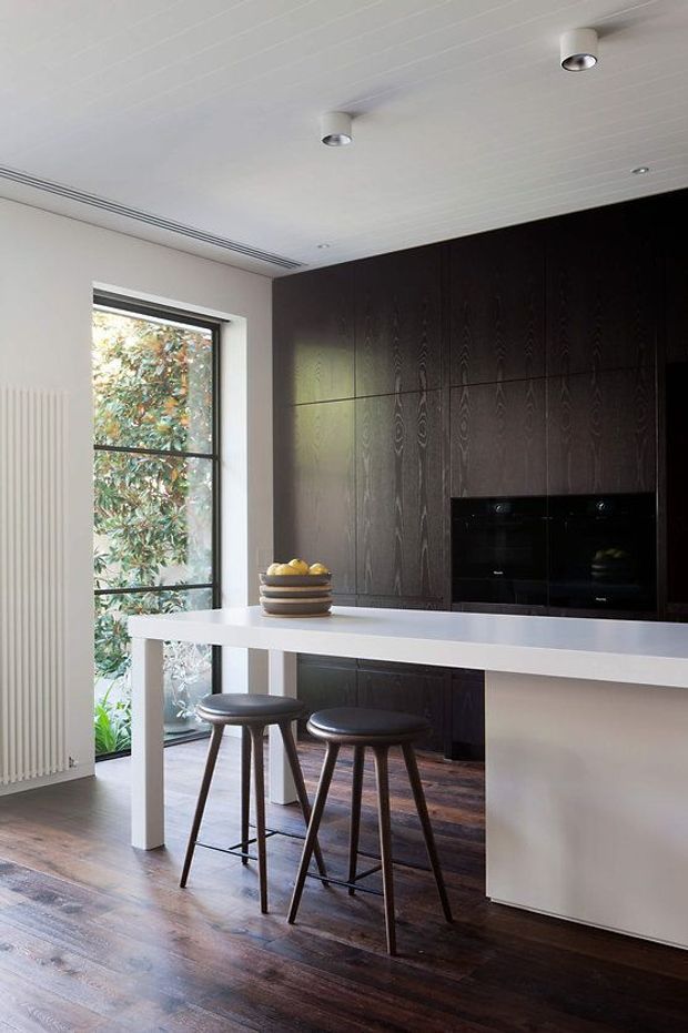 A refined contemporary kitchen with a long white island and integrated seating, dark timber full-height cabinetry, black built-in ovens, and dark timber flooring. A tall glazed door or window brings in natural light beside a slim white radiator. The ceiling has linear panel detailing and recessed spotlights, creating a clean, understated interior.