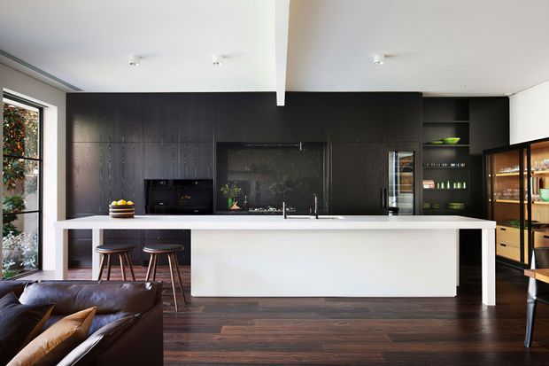 Contemporary open-plan kitchen viewed from the adjacent living area. A long white island with integrated sink and seating dominates the room, set against a full-height matte-black kitchen wall with dark cabinetry, black splashback, appliances, open shelving and a glazed display cabinet. Dark timber flooring, large black-framed glazing, recessed ceiling lights and a partial leather sofa create a refined, high-contrast interior.