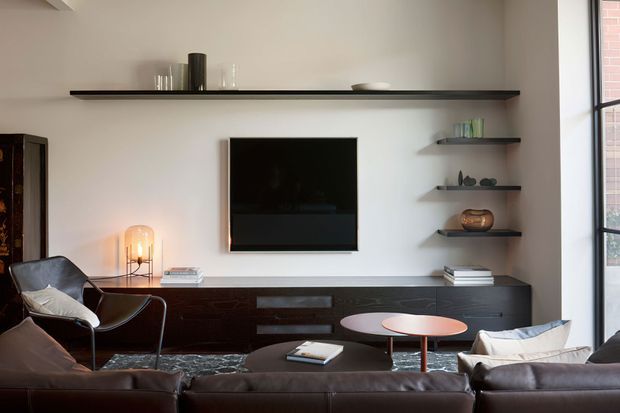 Contemporary living room with a dark leather sectional sofa and lounge chair arranged around a small round coffee table. A wall-mounted television is set above a long dark timber built-in media console, complemented by floating dark shelves displaying restrained decorative objects and books. A sculptural floor lamp, large black-framed window, neutral plaster walls, and an area rug complete the refined, warm interior.
