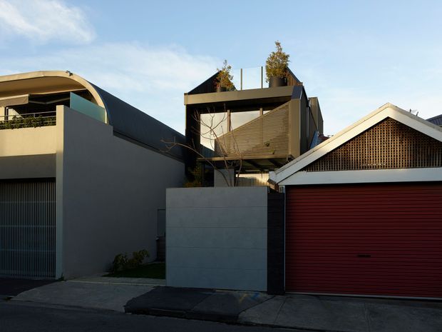 Street-facing view of a contemporary multi-level residential property, showing a dark rendered facade with curved roof forms, upper-level glazing and screening, rooftop planting, and a prominent detached-looking garage frontage with a red roller door and decorative timber ventilation screen. Concrete walls, driveway and pavement occupy the foreground.