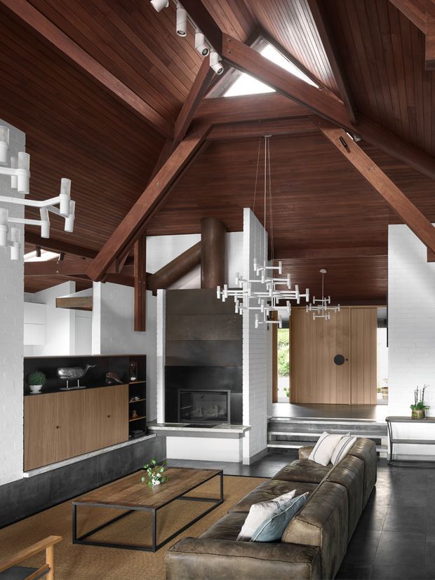 A dramatic open-plan living room with a soaring timber-lined pitched ceiling and exposed dark timber structural beams. Triangular skylights bring in natural light above a sculptural fireplace clad in dark stone or concrete. White brick walls, a built-in timber and black joinery unit, contemporary chandeliers, a low metal-framed coffee table, and a large leather sectional sofa create a refined contemporary interior. Steps lead to a timber front door at the rear.