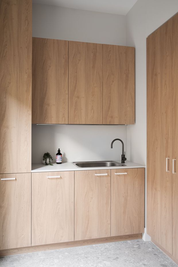 A compact, minimalist kitchenette with full-height light timber cabinetry, integrated upper and lower cupboards, a pale benchtop, stainless-steel sink, and dark curved mixer tap. Matching tall pantry-style cabinetry occupies the right side. The room has white walls and a large-format light grey stone-look tiled floor, creating a clean, understated fit-out.