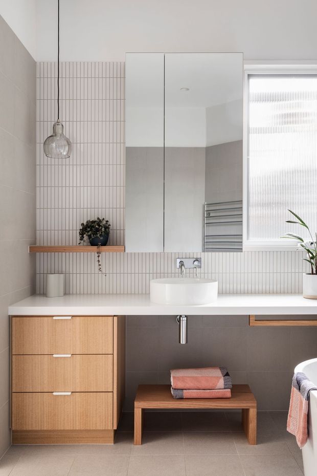 A bright, contemporary bathroom featuring a floating white vanity benchtop, integrated white vessel-style basin, wall-mounted tapware, mirrored cabinet, and warm timber cabinetry. Vertical slim white wall tiles form a feature backsplash, with large grey-beige tiles on the side wall and floor. A chrome heated towel rail, ribbed translucent window covering, pendant light, plants, and a small timber stool complete the calm, clean interior.