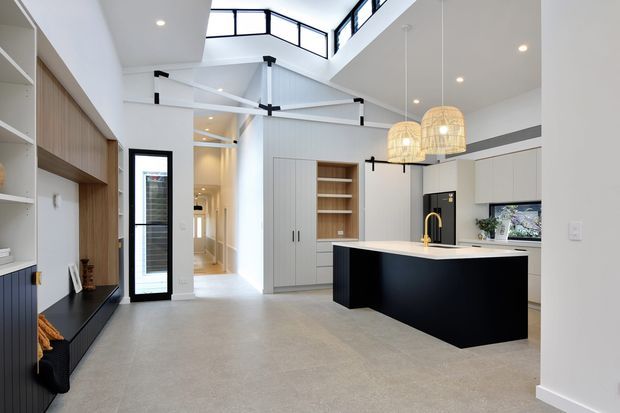 Bright open-plan contemporary kitchen and living area with a large black island, pale integrated cabinetry, timber-toned built-in shelving, pendant lights, and a double-height ceiling with clerestory glazing. A living/media joinery wall is visible on the left, while the kitchen occupies the central and right portions of the image.