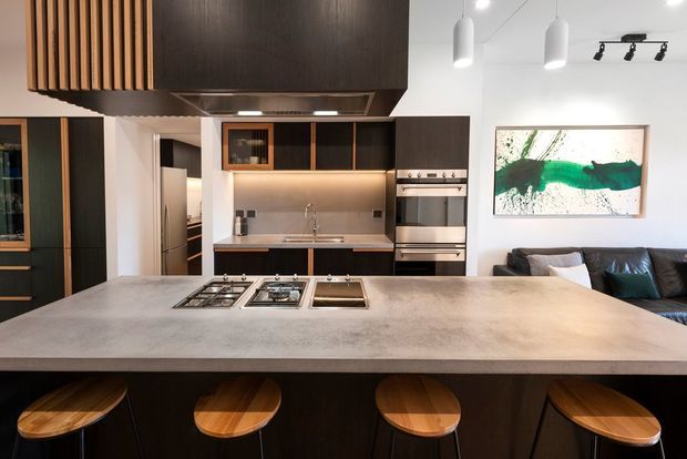 Contemporary open-plan kitchen viewed from behind a large island bench, featuring dark timber cabinetry, a pale stone or concrete-look benchtop, integrated cooktop and sink, pendant lighting, and timber bar stools. A living area with a sofa and abstract artwork is visible beyond the kitchen.