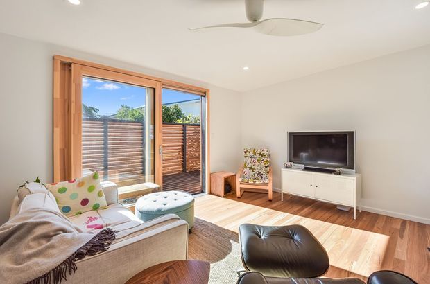 Bright, compact contemporary living room with a neutral upholstered sofa, colourful patterned cushions, a round ottoman, timber flooring, and a dark leather lounge chair. Large timber-framed sliding glass doors open to a fenced outdoor area, while a low white media console and wall-mounted television sit opposite the seating. Recessed ceiling lights and a modern ceiling fan provide lighting and ventilation.