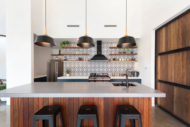 Contemporary kitchen with a large pale island and integrated sink, dark metal pendant lights, patterned geometric tile splashback, open timber shelving, stainless-steel appliances, and a richly stained vertical timber island front. A large timber-and-black framed sliding door is visible on the right.
