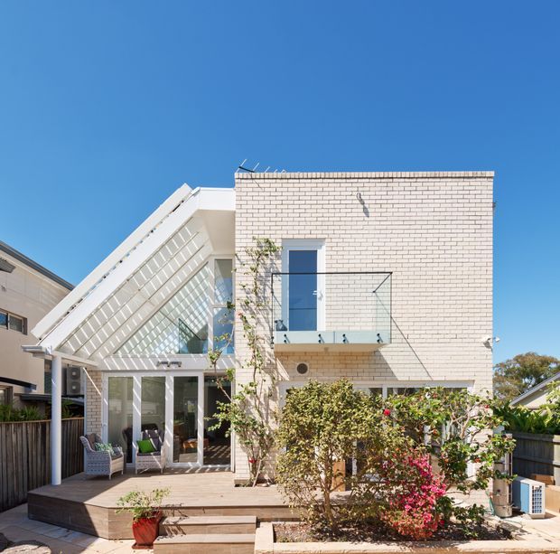 Exterior view of a contemporary multi-storey residence with a pale brick upper facade, a prominent steeply sloping glazed roof or clerestory structure, glass balcony balustrade, and large glazed doors opening onto a timber deck. Low garden planting and neighbouring homes surround the property under a clear blue sky.