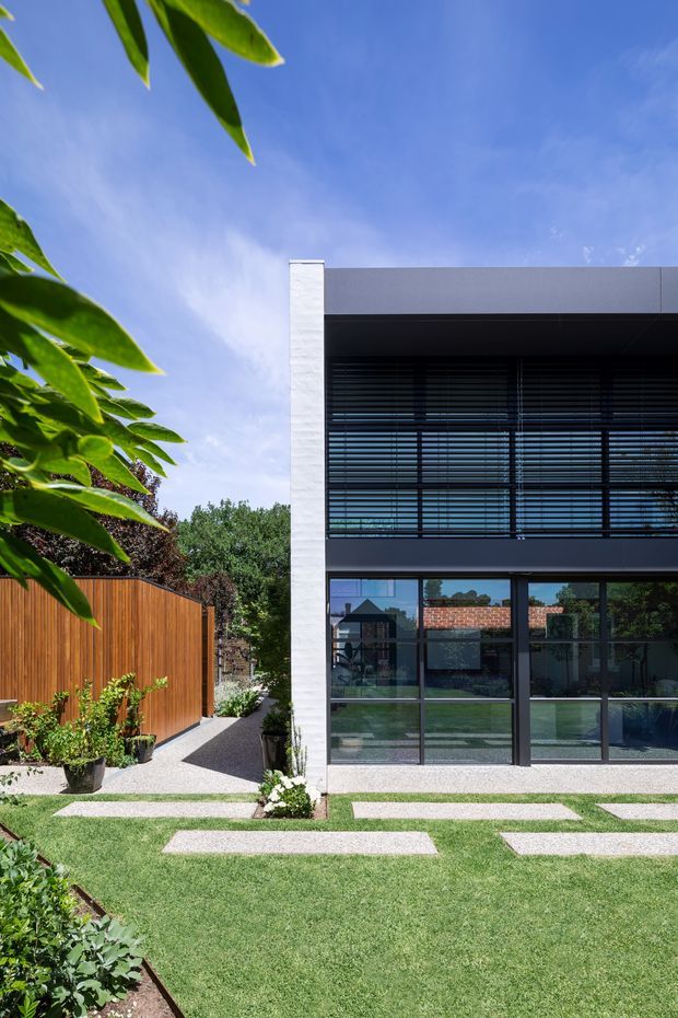 Exterior view of a modern two-storey residence with a crisp white rendered masonry side wall, dark framed floor-to-ceiling glazing, and horizontal external privacy sunshades. A manicured lawn with geometric pale stepping stones leads toward the building, alongside a warm timber privacy fence and landscaped planting. The image is brightly lit under a clear blue sky.