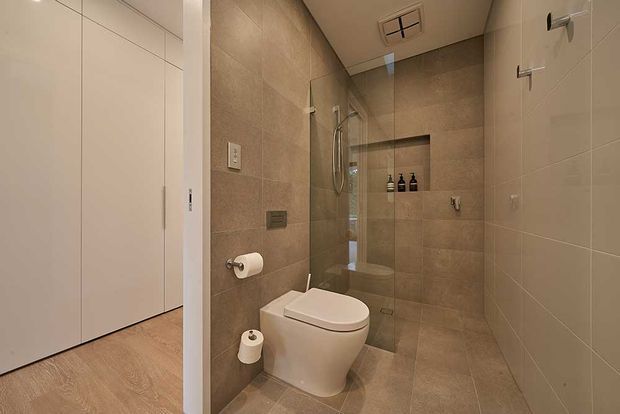 Compact contemporary bathroom with a wall-hung toilet, frameless glass walk-in shower, recessed shower niche, and tiled walls and floor in warm beige-grey tones. A white cabinetry or wardrobe area is visible through the open doorway on the left.