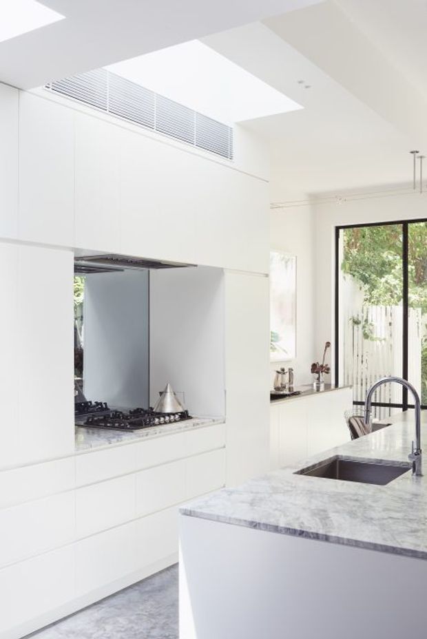 Bright, minimalist white kitchen with full-height handleless cabinetry, a concealed cooking niche with gas cooktop, marble-look island benchtop, and integrated dark sink with curved tap. A large skylight and high-level ventilation grille bring abundant natural light, while black-framed glazing opens toward a leafy outdoor area and dining setting.