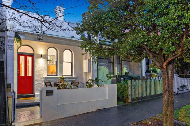Street-facing view of a compact attached residential terrace or townhouse facade, featuring rendered masonry walls, a vivid red entry door, arched windows, a pale metal pitched roof, a low rendered front boundary wall, and a small planted front garden behind a picket fence. A mature street tree partly obscures the facade.