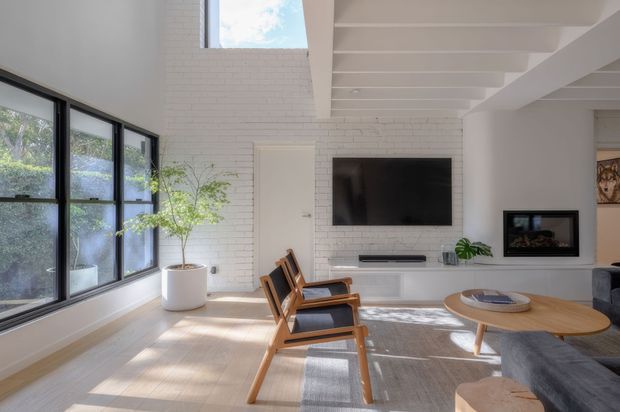 Bright contemporary living room with white-painted brick walls, exposed white ceiling beams, large black-framed windows, light timber flooring, and a wall-mounted television. Mid-century-inspired timber lounge chairs, a round timber coffee table, neutral rug, and indoor plants create a warm, airy seating area.