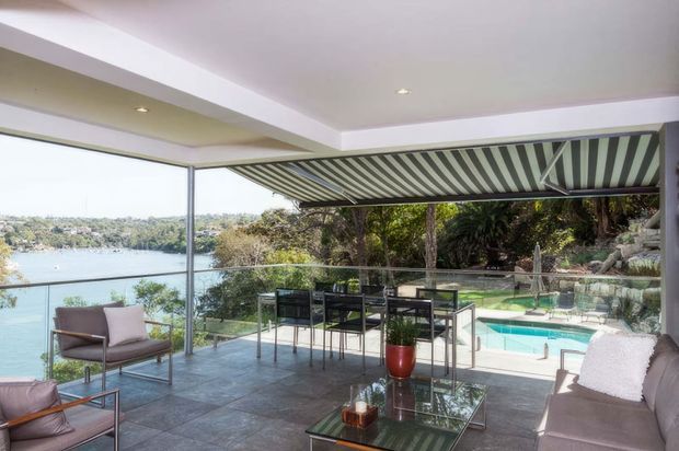 Covered outdoor terrace overlooking a waterfront landscape, furnished with an outdoor dining set, lounge seating, glass coffee table and a retractable striped awning. The terrace has large frameless-style glazed openings, dark tiled flooring, and direct views to a swimming pool and water beyond.