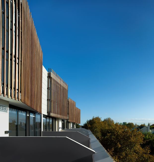 Exterior architectural photograph of a contemporary multi-storey building with long vertical timber-look facade fins, dark glazed ground-floor frontage, pale wall panels, and stepped dark parapets or terraces. The building recedes along the left side of the image, with trees and low residential buildings visible beyond under a clear blue sky.