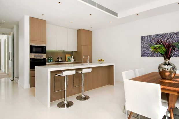 Bright open-plan contemporary kitchen and dining area with a white island, integrated appliances, light timber cabinetry, stainless-steel bar stools, and a timber dining table with white upholstered chairs. The space has a clean white palette, recessed lighting, and minimal detailing, with artwork and decorative objects adding subtle colour.