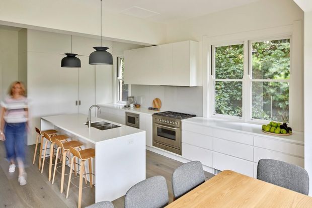 Bright, contemporary open-plan kitchen and dining area with a white island incorporating a sink and seating for several people, extensive white cabinetry, a stainless-steel range cooker, integrated appliances, timber bar stools, and a light timber dining table. Large windows overlook greenery, while black pendant lights provide contrast. A blurred person is present at the left edge.