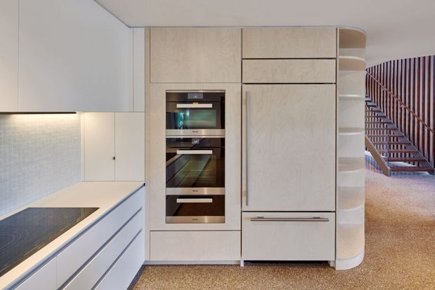 A contemporary minimalist kitchen with pale timber-look full-height cabinetry, integrated appliances, white drawers, and a light tiled splashback. A partially visible dark induction cooktop sits on the benchtop at left. The rounded end of the cabinetry forms open curved shelving beside a staircase with slim timber-toned balustrades. The space has a warm neutral palette and clean built-in detailing.