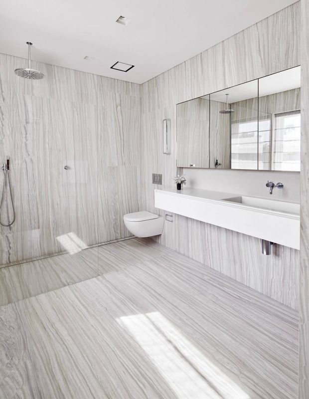 Bright, spacious contemporary bathroom with full-height pale grey-and-white veined stone-look wall and floor tiles. The room includes a walk-in shower with rain showerhead and handheld fitting, wall-hung toilet, and a long floating white double vanity with integrated basins, wall-mounted taps, and mirrored cabinetry. Minimal black and chrome fixtures, recessed ceiling lighting, and natural light create a clean, refined appearance.