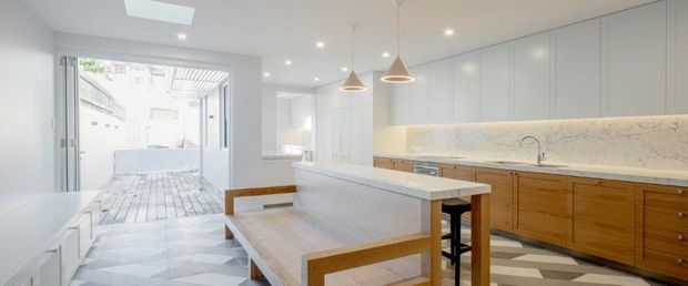 Bright contemporary kitchen with a large white island and integrated dining bench, warm timber lower cabinetry, pale stone benchtops and splashback, pendant lighting, and a light tiled floor. Full-height glazing or an open rear connection leads to an outdoor courtyard or deck.