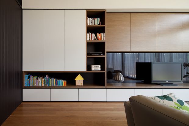 Contemporary living room focused on a large built-in entertainment and storage wall. The joinery combines matte white cabinets, warm timber veneer cupboards, dark open shelving, and a recessed television niche. A low sofa is visible in the foreground, with timber flooring and sheer curtains adding warmth and softness.