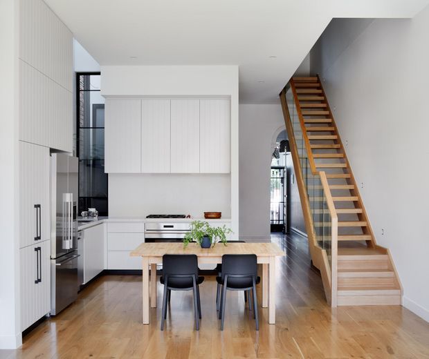 Bright open-plan kitchen and dining area with white flat-panel cabinetry, integrated stainless-steel appliances, a pale timber dining table, and two black dining chairs. A prominent open timber staircase with a simple balustrade rises along the right side, while dark timber flooring and full-height glazing contribute to the contemporary interior.
