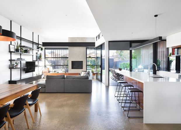 Bright open-plan contemporary home interior combining a kitchen, dining area and living room. The space features a long white island with integrated seating, dark-framed glazing, polished concrete flooring, open black shelving, a concrete fireplace wall, and access to an outdoor terrace or courtyard. Neutral furnishings and abundant glazing create an airy, refined atmosphere.