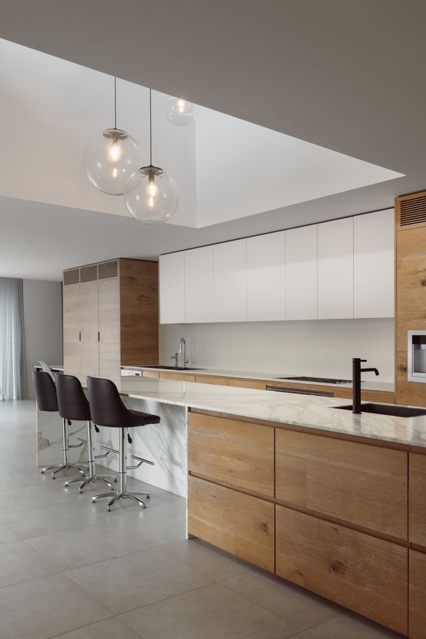 Contemporary open-plan kitchen with a large timber island, pale veined stone benchtops, white overhead cabinetry, integrated appliances, and a long breakfast bar with three dark upholstered stools. A sculptural recessed ceiling detail contains three clear globe pendant lights. The space has large-format light grey floor tiles, warm timber joinery, and a restrained neutral palette.