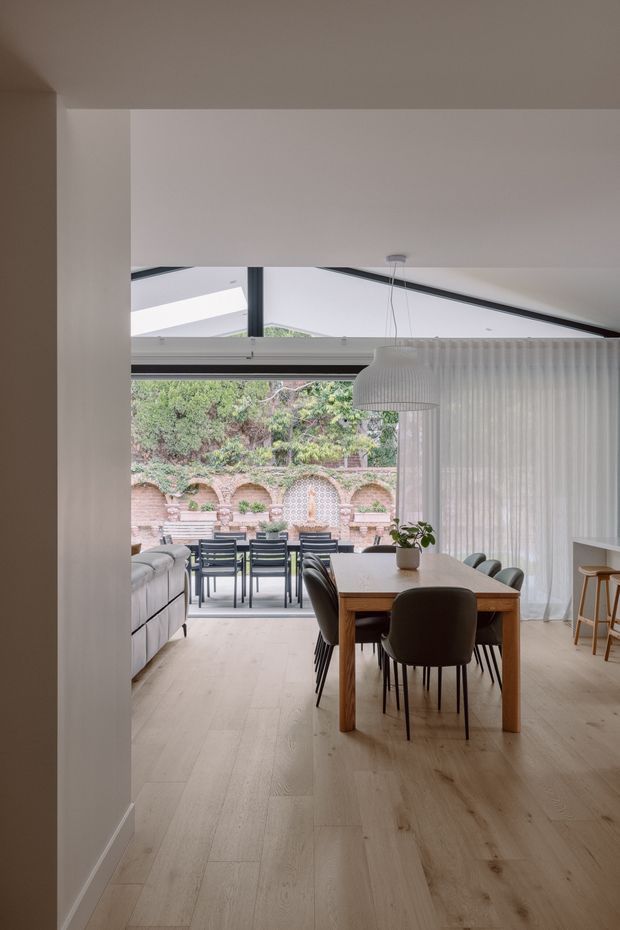 A bright, contemporary open-plan dining and living area viewed from an entry or hallway. The room has pale timber flooring, white walls and ceiling, a simple timber dining table with dark upholstered chairs, a white sofa, and a large ribbed pendant light. Full-height sheer curtains and broad glazed openings connect the interior to a covered outdoor terrace, with a landscaped brick garden wall visible beyond. A small kitchen or bar area with stools is partially visible at right.