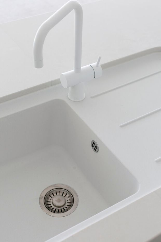 Close-up of a minimalist white kitchen sink with an integrated white benchtop and matching curved white mixer tap. The deep rectangular basin, stainless-steel waste fitting and clean seamless surfaces dominate the image.