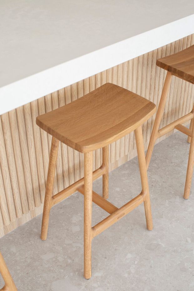Close-up interior detail of two light timber bar stools positioned at a white kitchen or bar counter. The stools have simple rounded rectangular seats, tapered legs, and horizontal footrests. Behind them is a vertical light-wood slat wall, while the floor appears to be a pale mottled concrete or stone-like finish. The image has a clean, warm, minimalist aesthetic.