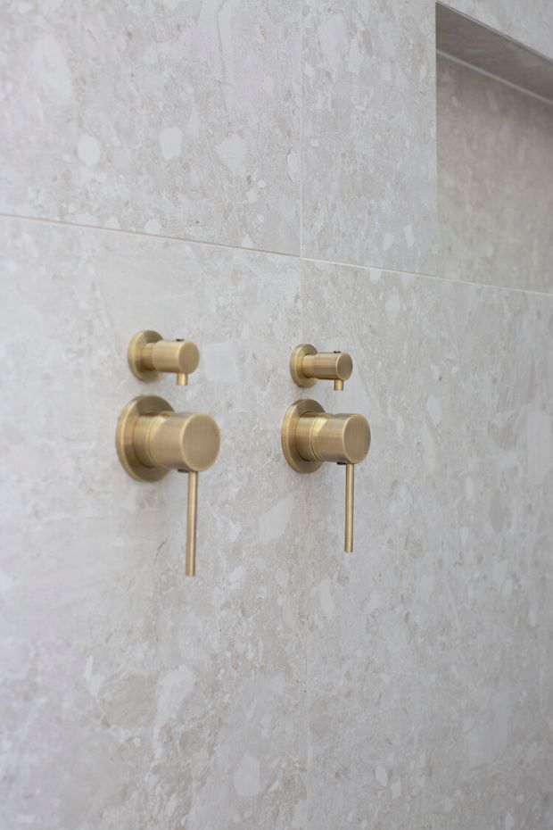 Close-up of a refined bathroom wall featuring two sets of brushed brass/gold shower controls mounted on large-format pale grey stone-look tiles. A mirror or reflective surface is visible at the upper right, reinforcing the bathroom setting. The composition is minimalist and detail-focused, with soft neutral tones and metallic accents.