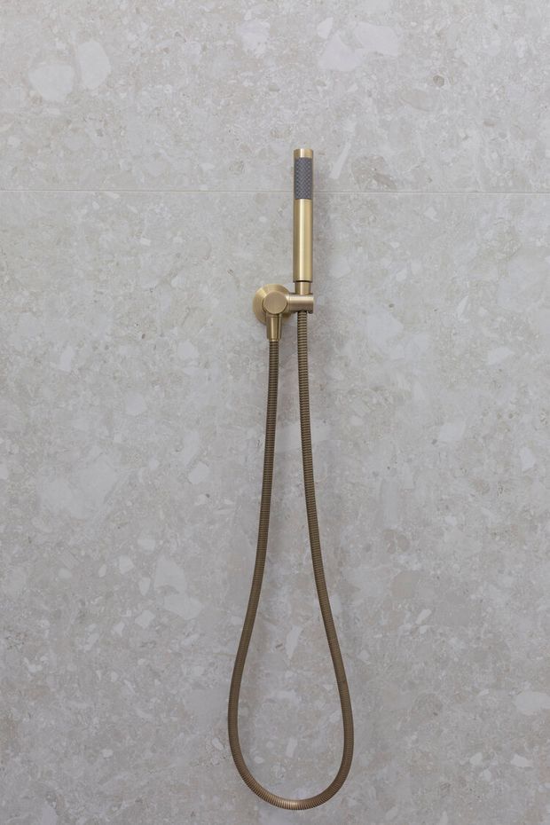 Close-up of a wall-mounted handheld shower set with a cylindrical brushed brass-gold handset, matching round control bracket, and flexible metal hose against large-format pale grey stone-look bathroom tiles.