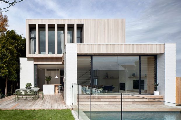 Exterior view of a contemporary two-storey residence with a rectilinear form, pale vertical timber-like cladding, white masonry walls, tall dark-framed windows, and extensive ground-floor glazing. A timber deck, glass balustrade, and swimming pool occupy the foreground, with glimpses of a bright open-plan interior and outdoor seating beyond.