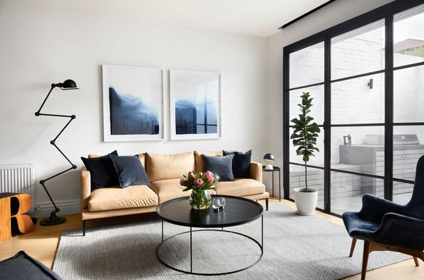 Bright contemporary living room with a tan sofa, navy cushions, sculptural black floor lamp, round black coffee table, textured light rug, and a dark blue armchair. Large black-framed grid glazing opens visually to a light-filled outdoor area, while abstract blue artwork and a potted tree add restrained accents.