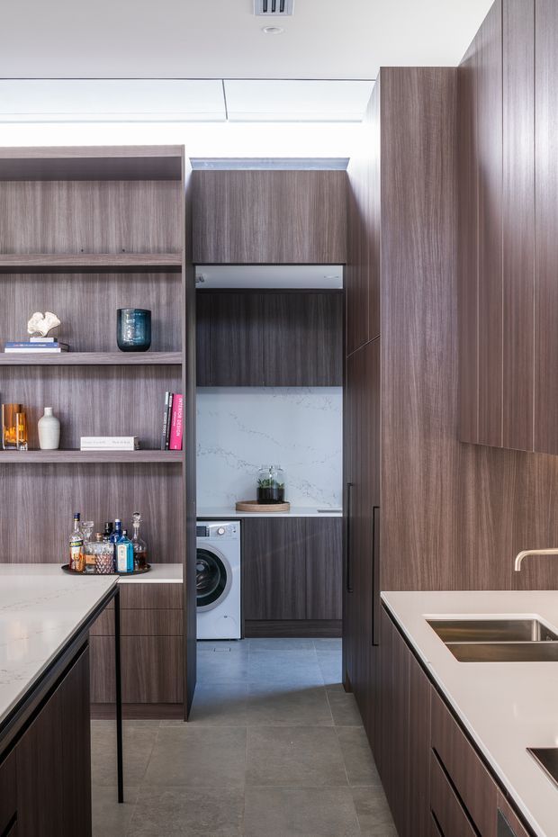 A narrow contemporary kitchen viewed toward an adjoining laundry area. Floor-to-ceiling dark timber-grain cabinetry and open shelving line both sides, with pale stone benchtops, an integrated stainless-steel sink and tap, and a marble-look stone splashback in the laundry nook. Grey large-format floor tiles, concealed appliances, and minimal decor create a streamlined, space-efficient fit-out.