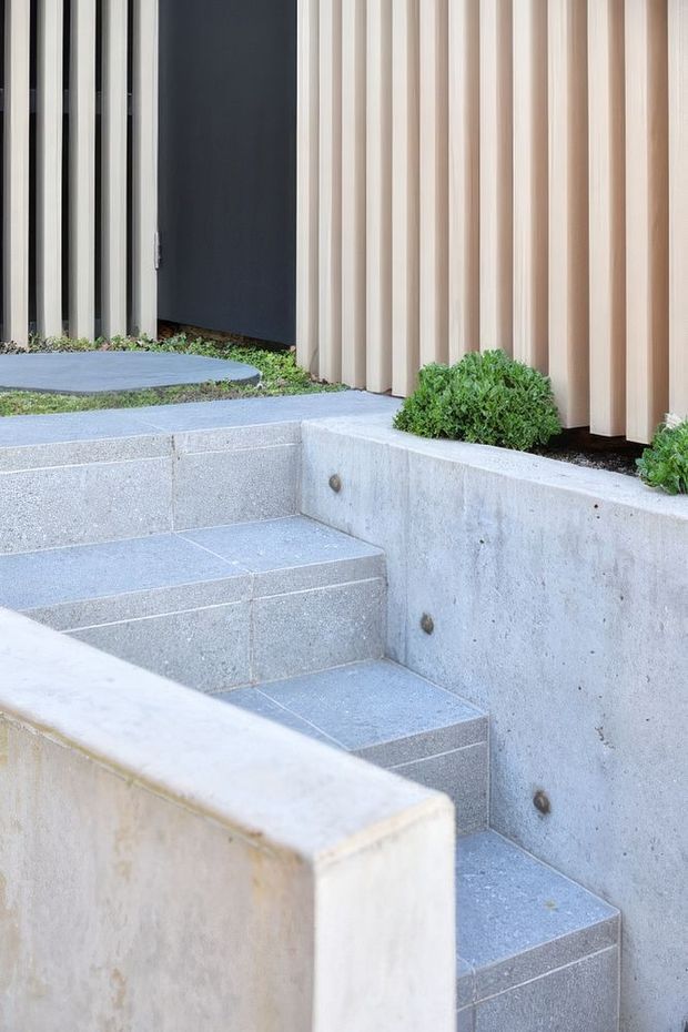 An exterior entry approach with broad grey tiled steps set between pale concrete retaining walls. The steps lead toward a dark entry door framed by vertical black and light timber-toned facade elements, with low green planting integrated along the edges. The composition is clean-lined and contemporary.