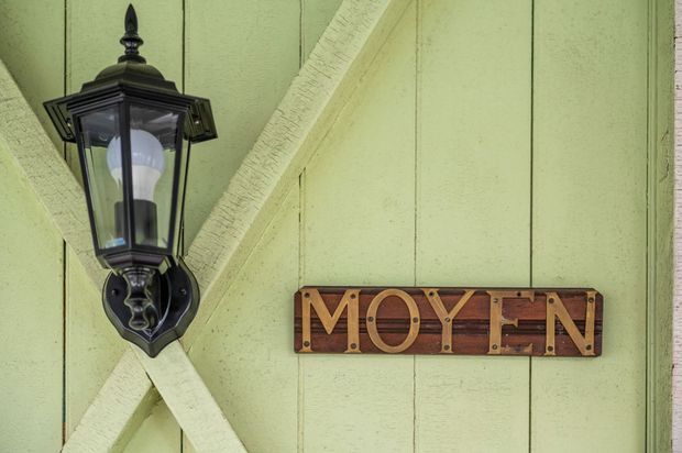 Close-up of a pale green painted timber-clad exterior wall with diagonal structural bracing, a black traditional wall-mounted lantern, and a rustic wooden sign reading “MOYTEN.”