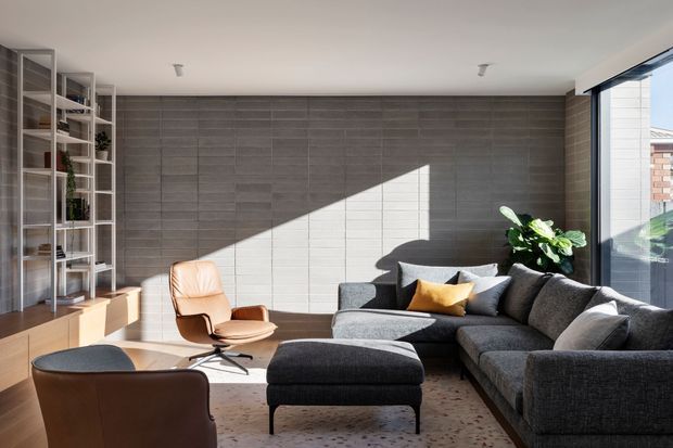 Contemporary living room with a large charcoal sectional sofa and ottoman, a tan leather lounge chair, timber built-in cabinetry and open shelving, and a broad grey tiled feature wall. Strong sunlight enters through expansive corner glazing, creating a bright diagonal highlight across the wall. The room has a calm, refined, modern feel with indoor planting and restrained furnishings.