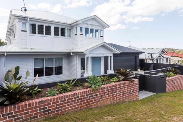 Street-facing exterior of a two-storey suburban home with white weatherboard cladding, a pitched gabled roof, louvred upper-level windows, dark garage door, and a landscaped front garden enclosed by a red brick retaining wall.