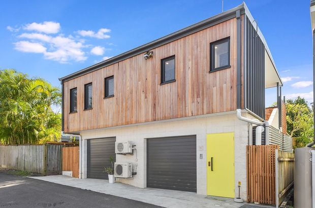 Exterior view of a compact contemporary multi-level building with a white masonry ground floor containing two dark roller garage doors and a bright yellow pedestrian door. The upper level is clad in warm vertical timber boards with dark-framed windows and a dark ribbed metal side wall. Timber fencing, exposed services, a narrow driveway, and tropical landscaping are visible under a clear blue sky.