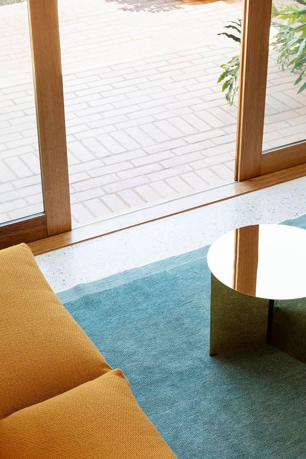 A bright, refined living area viewed from above, featuring a mustard-gold upholstered sofa, a blue-green area rug, a small round white tabletop with dark base, and warm timber-framed floor-to-ceiling glazing. A pale brick-paved outdoor area and greenery are visible beyond the windows.